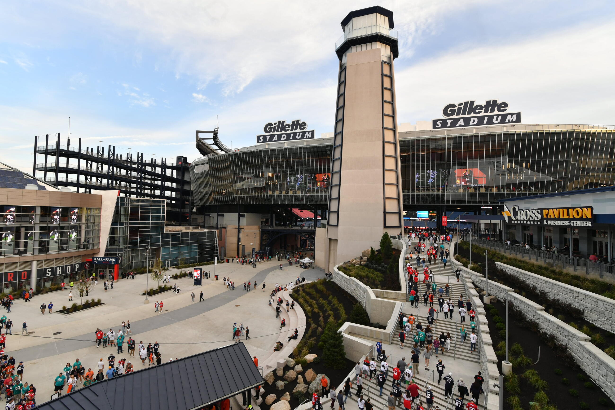 a view of gillette stadium from the outside with lots of people walking on the stairs and in the open plaza area