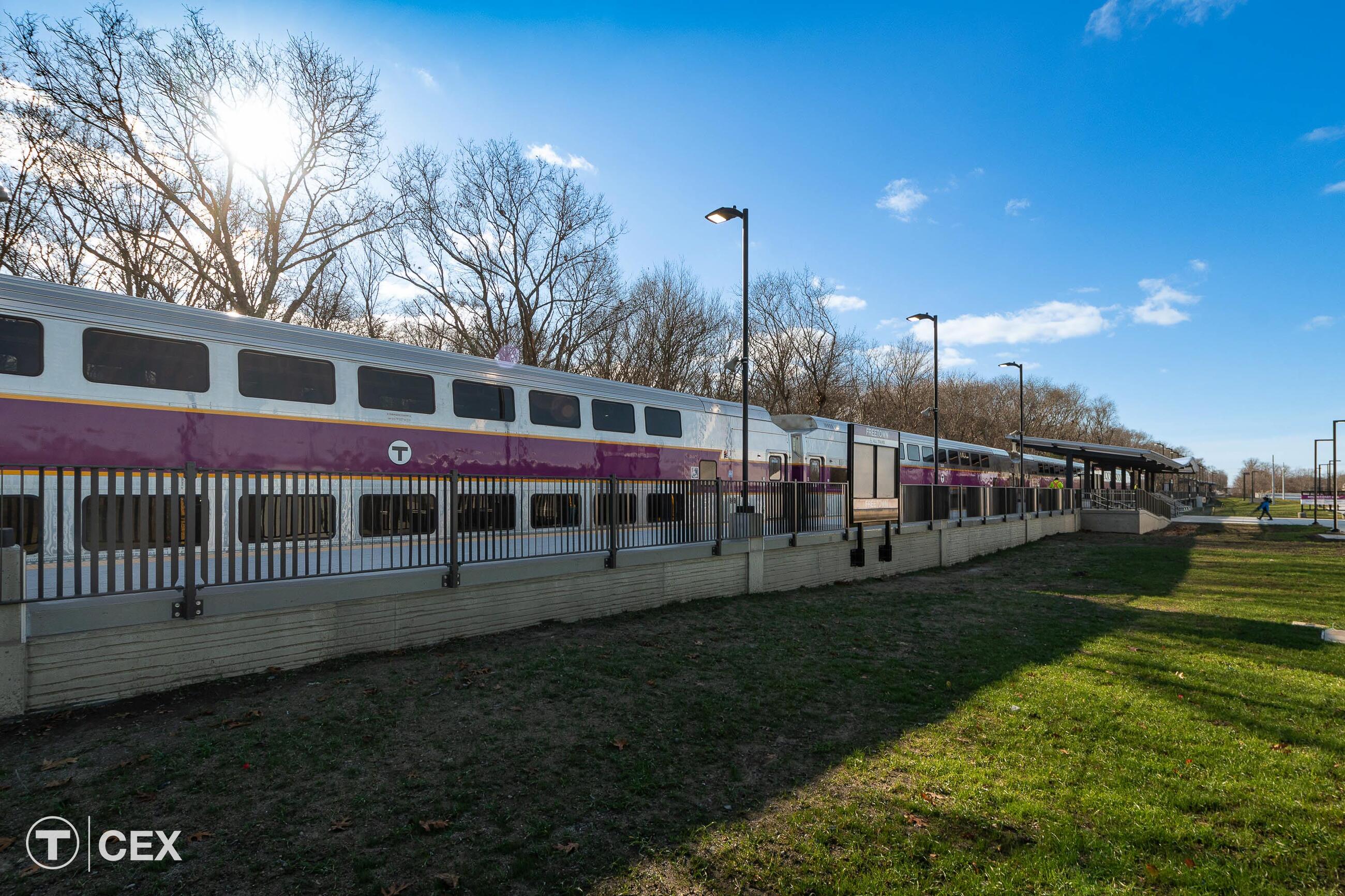 Silver train with purple stripe sits on a track. There is green grass in the foreground.