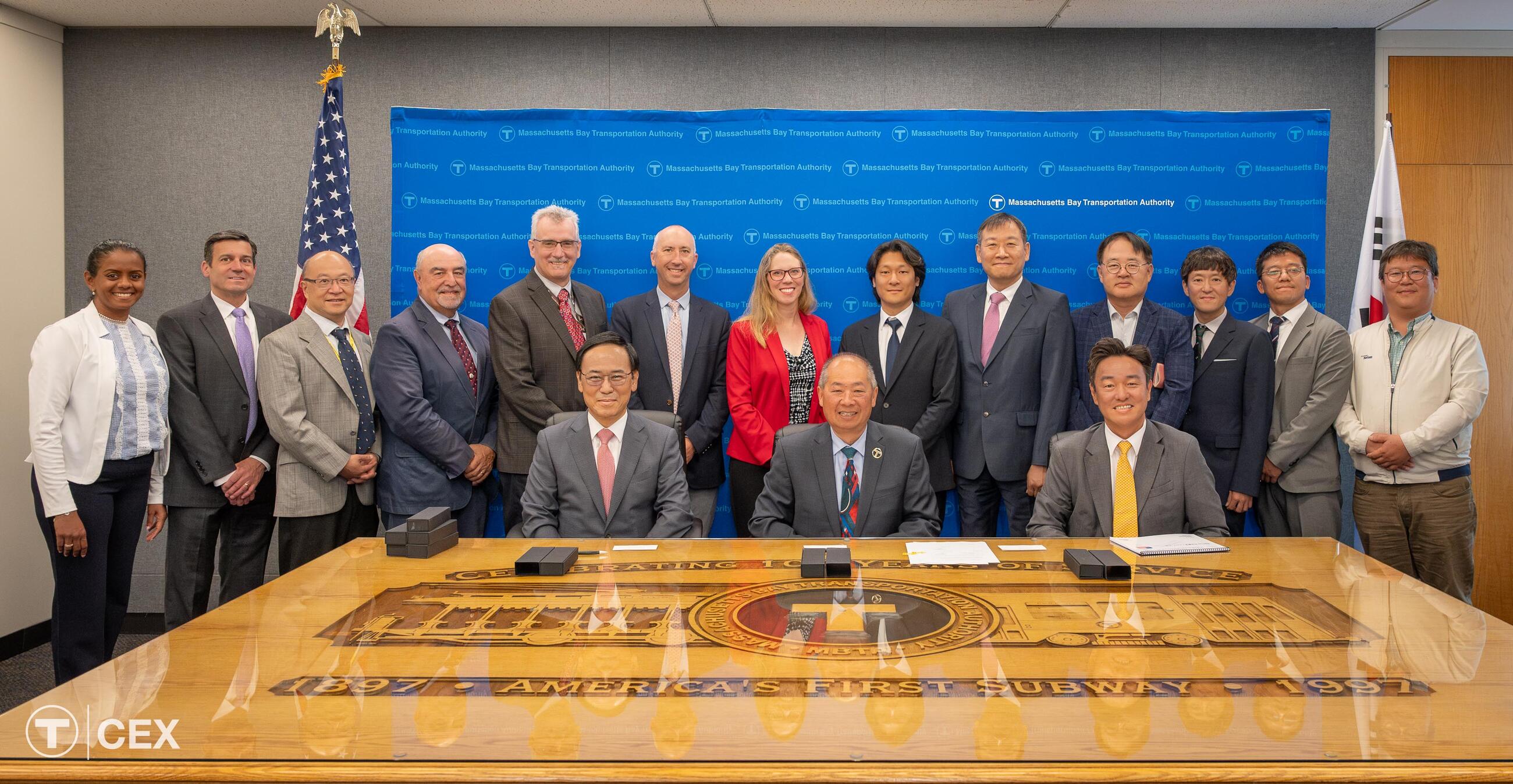 13 individuals stand behind 3 seated individuals, all in front of a blue background and behind a large brown table.