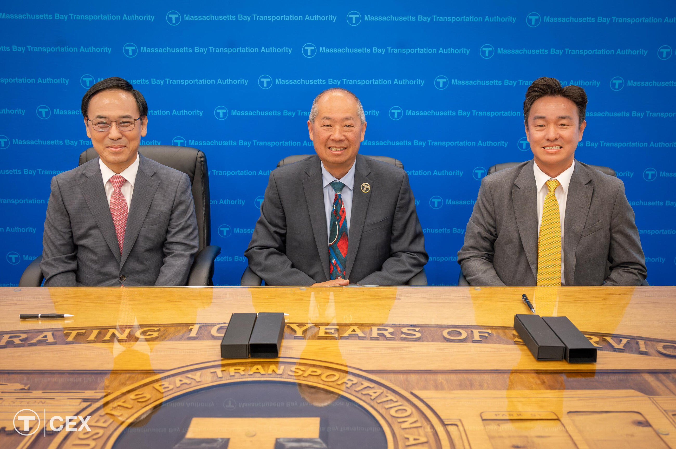 Three men sit in black chairs behind a big brown table and in front of a blue background.