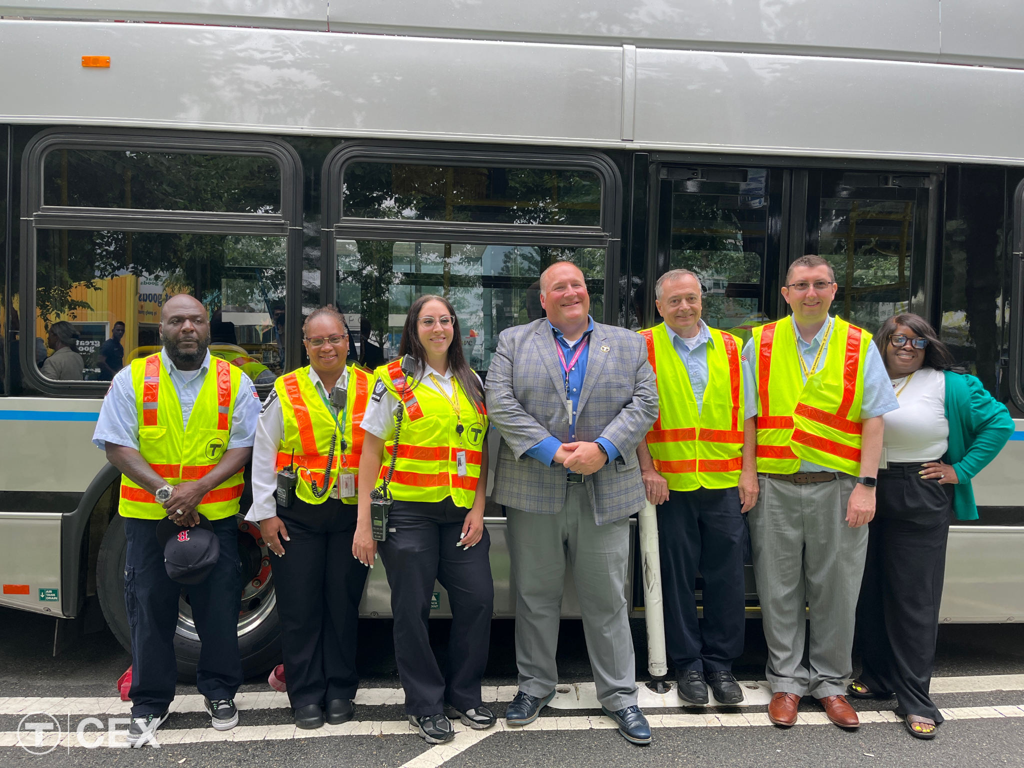 Seven people stand in front of a silver bus together in one line