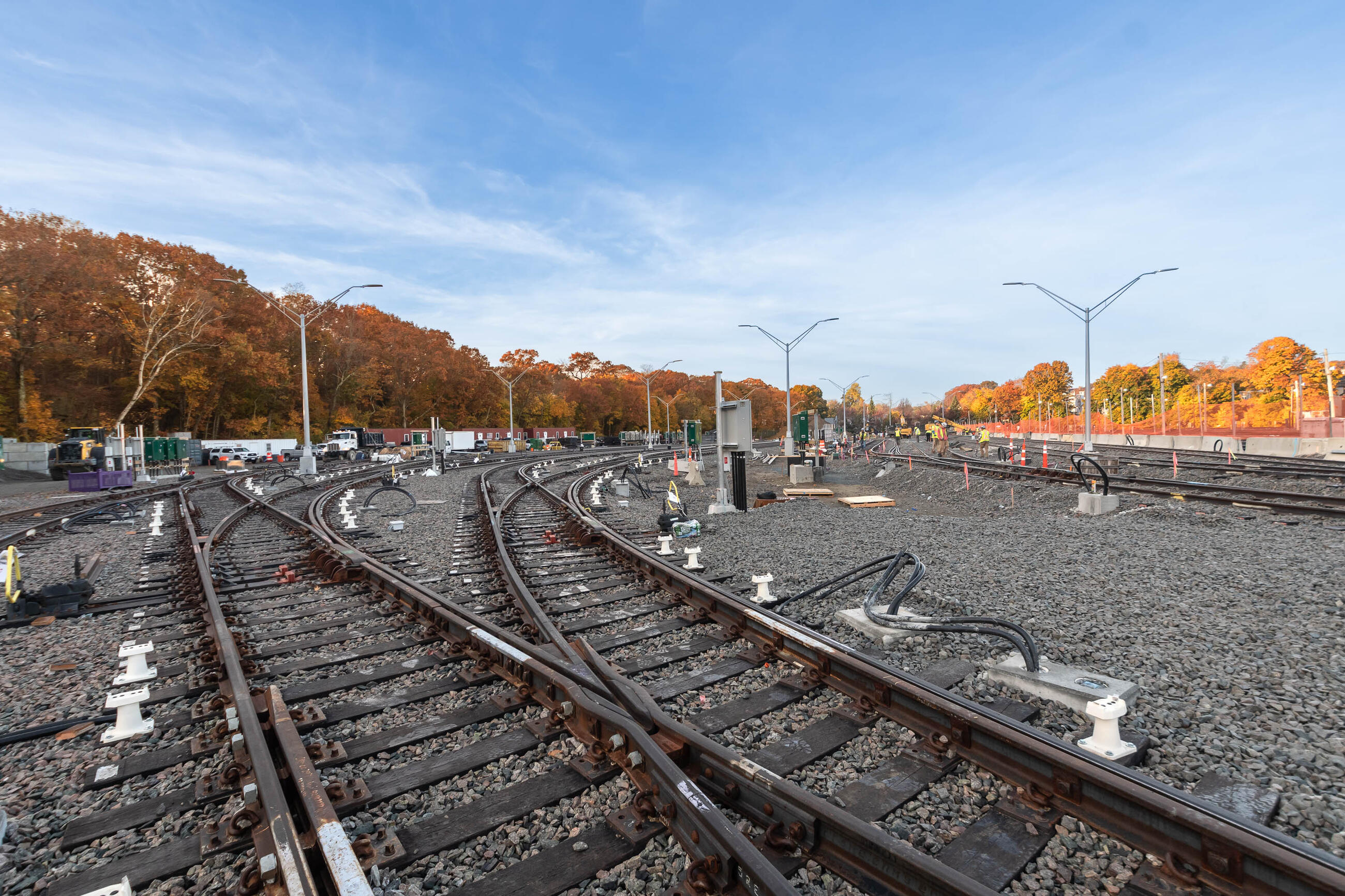 Several train tracks under construction converge in the work zone. Some construction trucks and workers are in the distance, with fall foliage in the background.