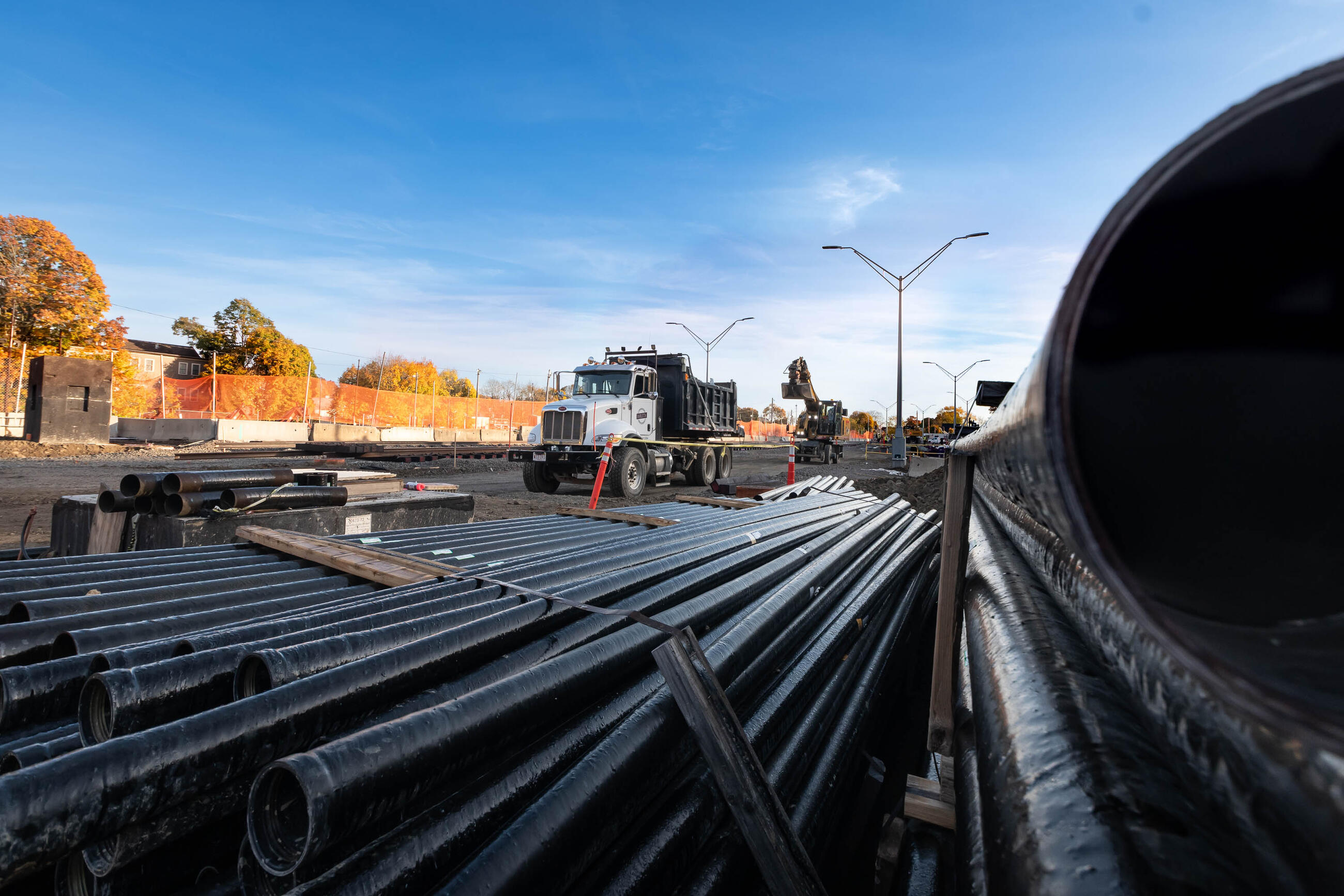 A close-up view of a pile of pipes waiting for installation, with a construction truck and fall foliage in the background