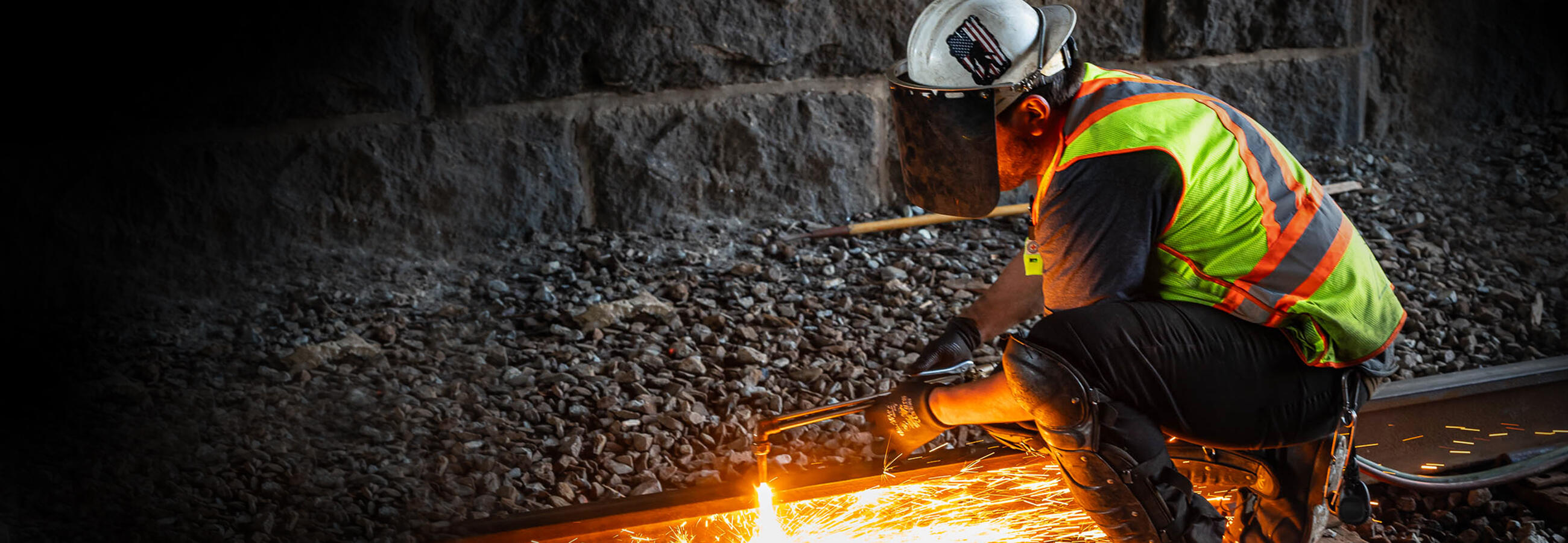 A welder wearing a yellow safety vest works on tracks. Sparks appear from the welding.