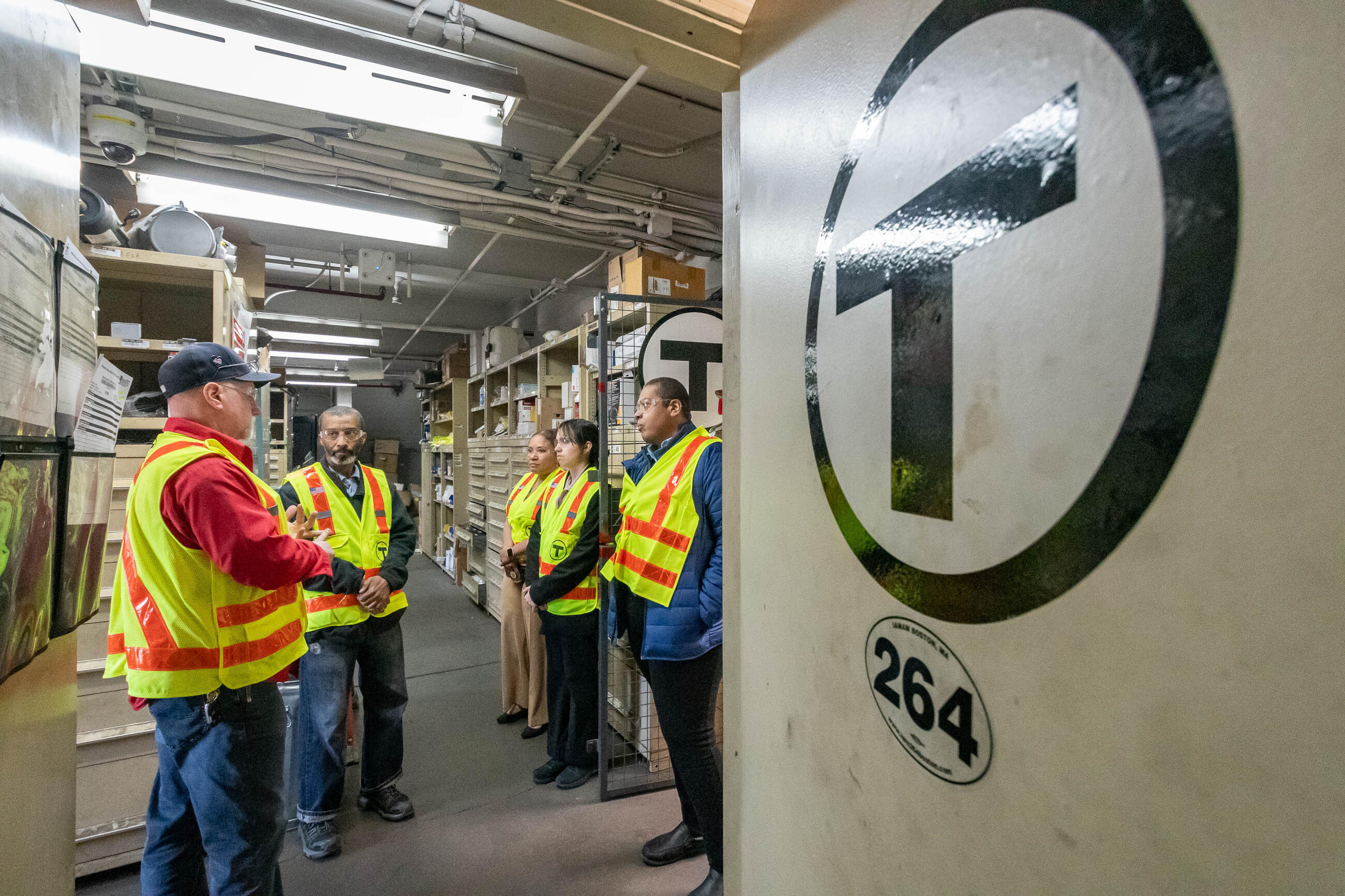 Five different people in yellow construction vests and safety glasses standing in a storage room talking, next to a big MBTA logo on the wall.