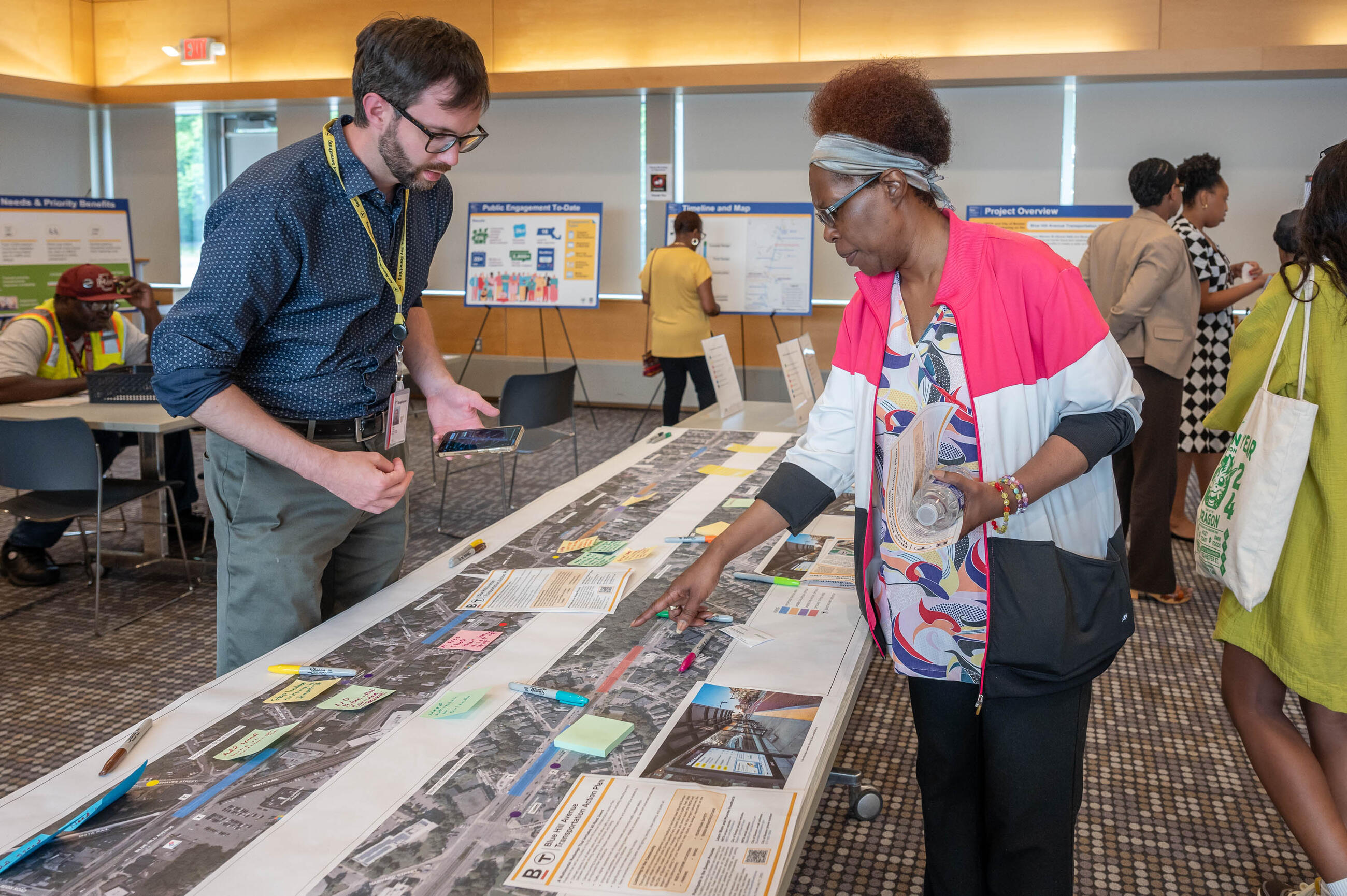 Two people standing on either side of table in a meeting room, pointing at and discussing a map that covers the table. A few other people in the room look at presentations on easels in the background.