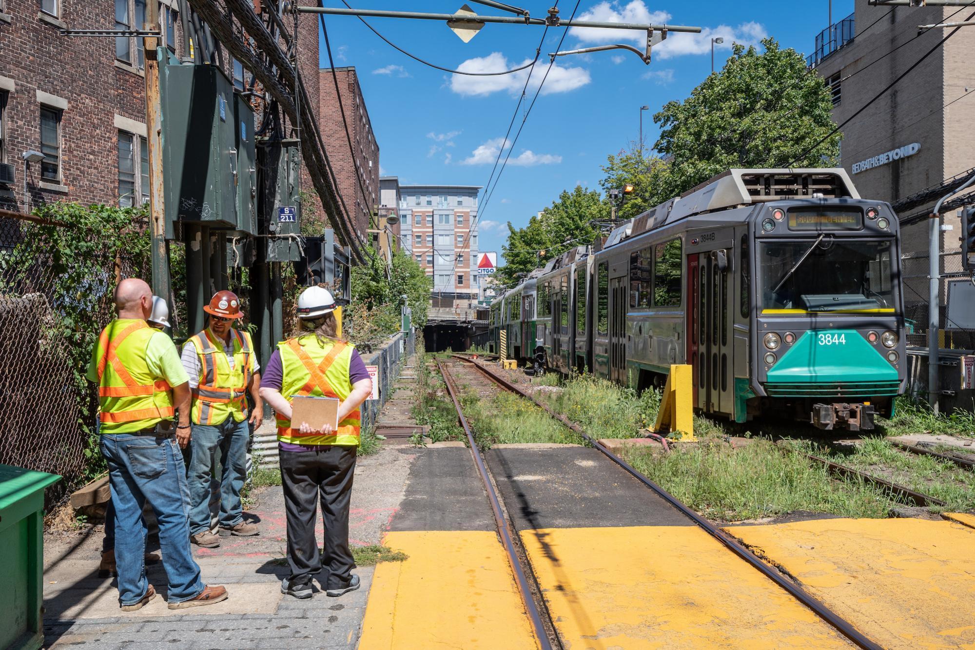 A three-person construction crew stands on the left as a Green Line D train exists the Fenway Portal and pulls into Fenway Station.
