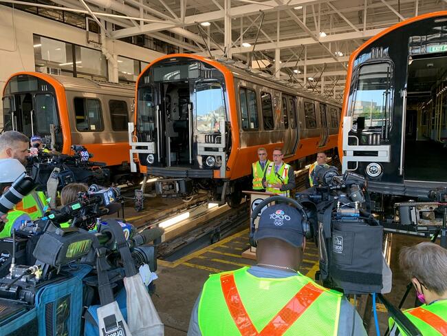 MBTA General Manager Steve Poftak updates members of the media standing at a podium in front of 3 new Orange Line subway cars.