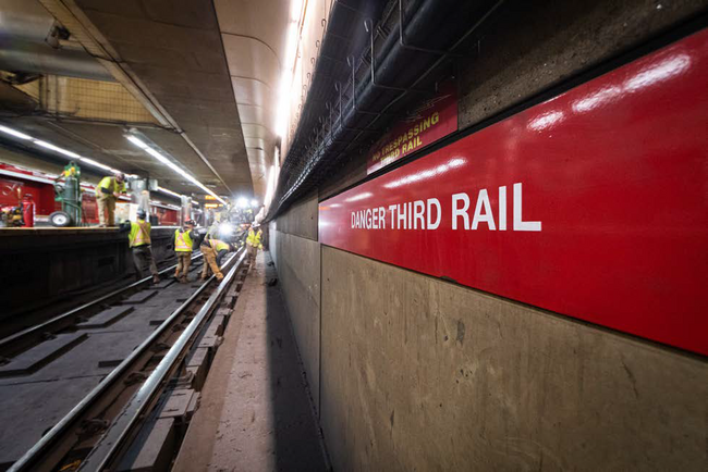A “Danger Third Rail” sign on the Red Line right of way with several personnel working in the background