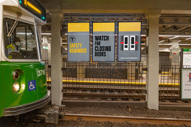Above Green Line tracks, in front of a train, digital signage shows a safety reminder to “watch for closing doors”