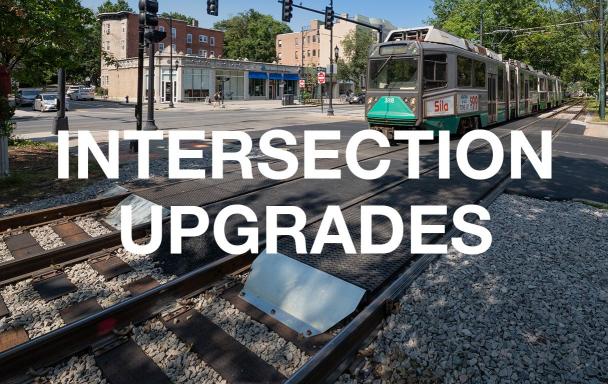 A closeup of an upgraded intersection, with a Green Line C train approaching. White text overlaid reads "Intersection Upgrades."