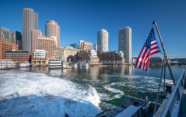 A photo of Rowes Wharf, taken from the ferry across the water. The water is white from the wake of the ferry moving in the water. A United States flag is flying at an angle from the side of the boat, and the sky is a bright clear blue 