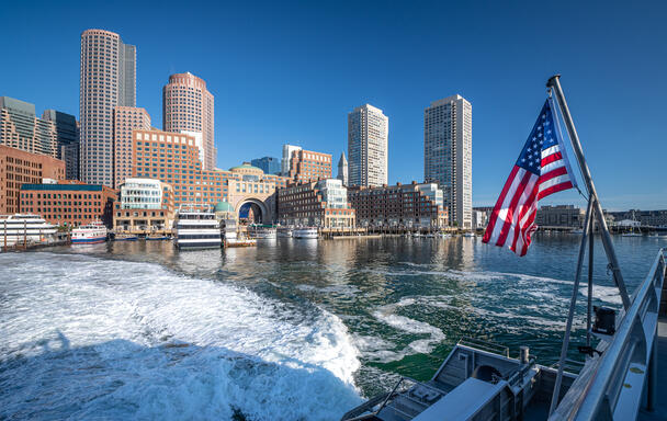A photo of Rowes Wharf, taken from the ferry across the water. The water is white from the wake of the ferry moving in the water. A United States flag is flying at an angle from the side of the boat, and the sky is a bright clear blue