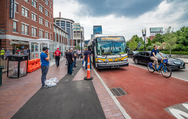 Riders wait in line to board a bus in front of a brick building labeled Registry of Motor Vehicles. The bus, which has a sign spelling out FACE COVERING REQUIRED displayed, is at a Haymarket Station bus stop on Surface Street. Cars and a bicyclist pass to the right of the bus.