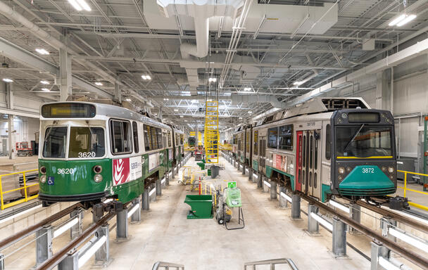 A photo of green line trolley cars at a GLX testing facility. The train on the left is new, and the one on the right is old. They are both elevated on rails inside of a brightly lit warehouse