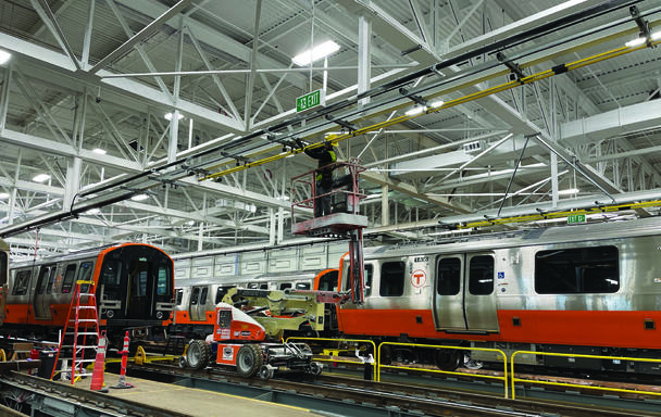 a crew member uses a lift to work on something near the ceiling inside the wellington vehicle maintenance facility, with new orange line train cars on the ground next to him