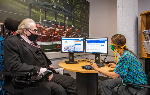 A woman sitting at a desk at the MBTA's Mobility Center assists a man in a motorized wheelchair with planning a trip on the computer. They are both looking at the computer screens as the woman moves a mouse.