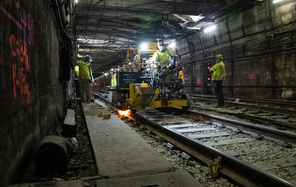 A construction worker drives a piece of machinery over train tracks in a tunnel on the Blue Line. Other workers to the left and the right of the tracks watch as the machinery drives by.