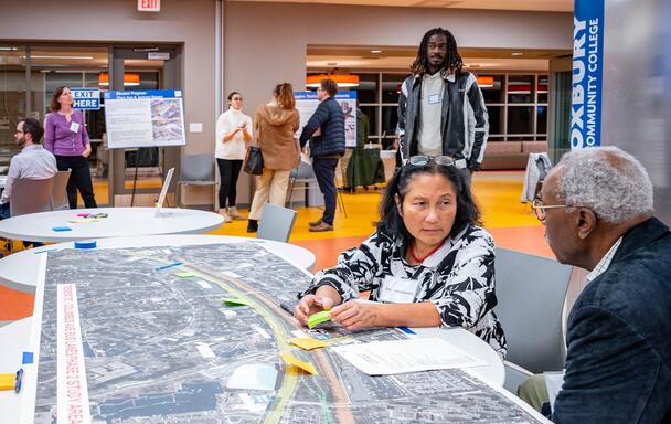 Two community members discussing a map laid out on a table with other members standing and talking in the background